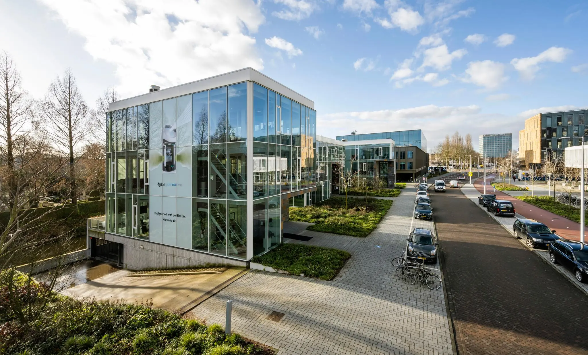 Modern kantoorgebouw aan de Fred. Roeskestraat met glazen gevel en een Dyson-reclame, gelegen langs een straat met geparkeerde auto's en fietsen.